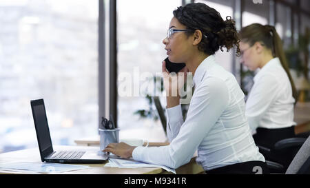 Beautiful mixed-race lady sitting at office and talking on phone with customer Stock Photo