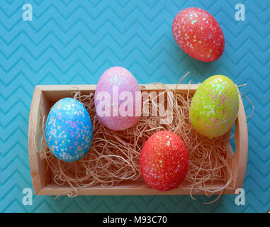 five colorful Easter Eggs in a wooden box with nesting material inside on a pastel blue background Stock Photo