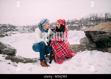 Young couple in hats and scarfs with dog wrapped in red plaid during winter walk next to frozen lake and rocks. Stock Photo
