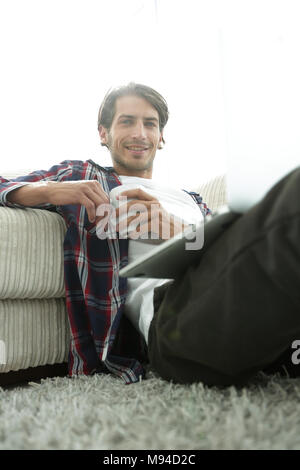 young man with laptop holding a cup sitting on the floor near the sofa Stock Photo