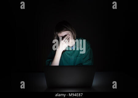 Tired and exhausted young woman rubs eyes at laptop pc late in the evening. Portrait of depressed female student or worker sitting in front of compute Stock Photo