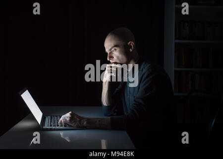 Attentive and concentrated young man at laptop pc late in the evening. Portrait of serious male student or worker sitting in front of computer screen  Stock Photo