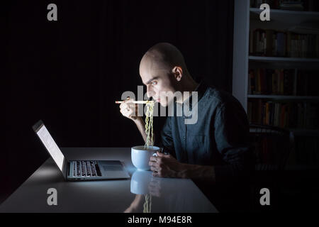 Young male at laptop computer eats instant ramen noodles with chopsticks late in the evening. Man working or studying online overtime at night has fas Stock Photo