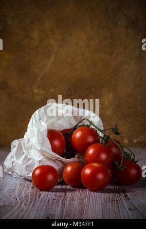 Vertical photo with few twigs with small red cherry tomatoes. Vegetable is in white paper bag which is placed on wooden vintage board with worn surfac Stock Photo