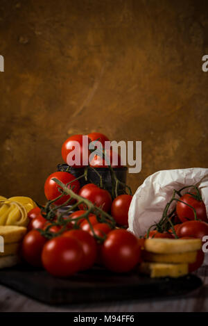 Vertical photo with small red cherry tomatoes which are on green twig and placed in vintage metal worn can. Tagliatelle and bruscheta rings are placed Stock Photo