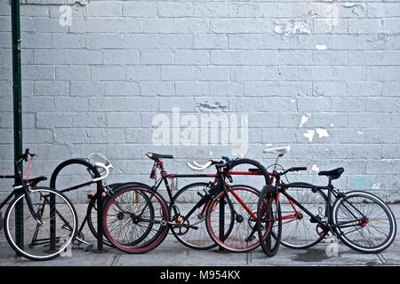 Queens, New York, USA: Bicycles chained to sign posts and curbside bike stands on a sidewalk, with a grey painted wall in the background. Stock Photo