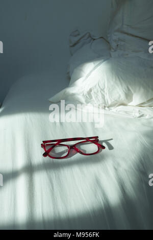 close up of ladie’s red reading glasses lying face down on a sunlight bed with white sheets and pillow Stock Photo