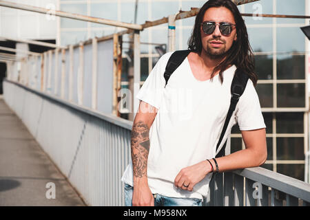 Young attractive bearded man with tattoos and long hair wearing sunglasses, white shirt and backpack posing on urban background Stock Photo