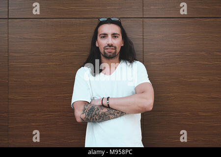 Beautiful long haired man with beard and tattoos posing on a wood wall with crossed arms while looking to camera, looking to right. Copy space Stock Photo