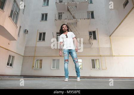 Low angle shot of Stylish man posing on building background. Male looking to right while fixing sunglasses with tatood hand. Stock Photo