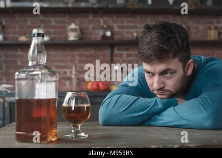 Young man alcohol addiction problems sitting at table looking at whiskey bottle and glass fighting with addiction Stock Photo