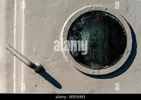 Dark and murky tempered glass porthole on white ferry door from with handle casting an afternoon shadow and the porthole casting a crescent shadow Stock Photo