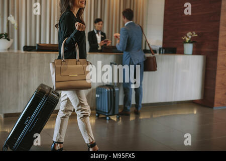 Female guest walks inside a hotel lobby with people in the background at reception counter. Woman arriving at hotel. Stock Photo