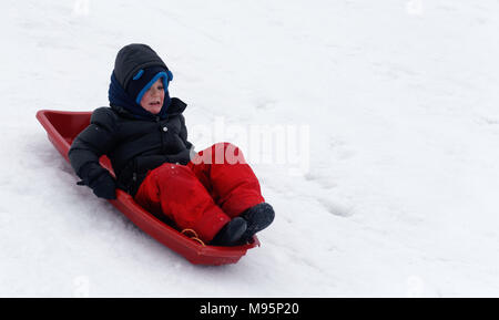 A little boy (5 yr old) looking scared as he sledges down a steep icy slope Stock Photo