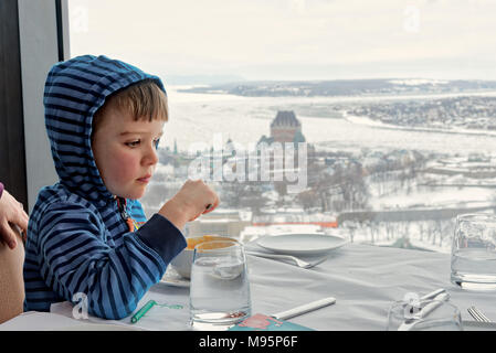 A five year old boy eating in the Ciel! rotating restaurant in Quebec City, with the Chateau Frontenac and the frozen St Lawrence beyond. Stock Photo