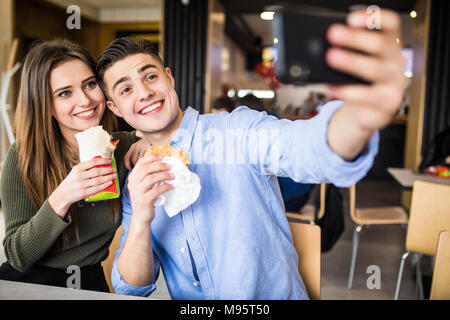 Happy couple eating burger and doner taking selfie together in fast food Stock Photo