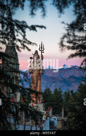 The tall golden Shiva staute of Khajjiar during the sunset with snow peaks in the background and peeking through the trees Stock Photo