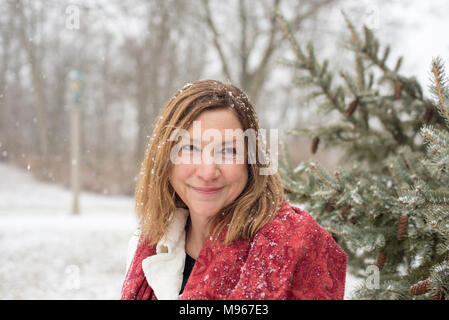 smiling woman standing by snow covered pine tree in park with magical fresh snow falling Stock Photo