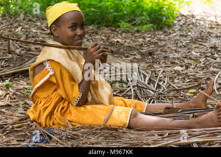 Shy young African girl stripping dried palms leaves to make into brooms. Stock Photo