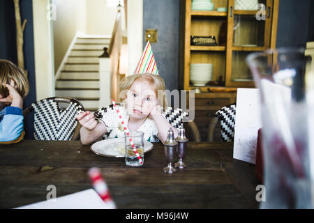 Girl sitting at table eating cake with a party hat on Stock Photo