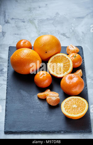 High angle still life view of sliced ruby orange and tangerine arranged together on the right side of black stone cutting board over white textured ba Stock Photo