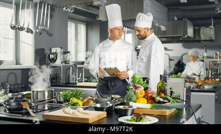Two Famous Chefs Exchange Ideas About Video Blog Recipe they Watching on Tablet. They are Working in Big Restaurant Kitchen. Stock Photo
