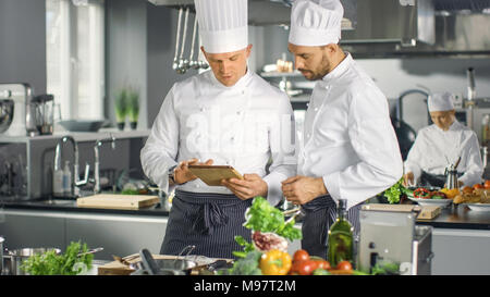 Two Famous Chefs Exchange Ideas About Video Blog Recipe they Watching on Tablet. They are Working in Big Restaurant Kitchen. Stock Photo
