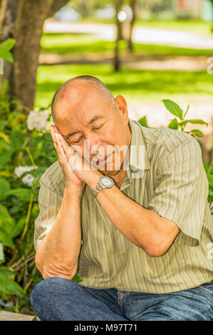 Outdoor view of old man sits on bench, enjoying the nature and having a good rest with both hand in his face, pretending sleeping. All problems left behind Stock Photo