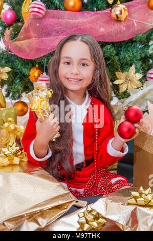 Close up of smiling girl wearing a red santa costume and holding two christmas ball in one hand and two gifts in her other hand, with a Christmas tree behind, christmas concept Stock Photo