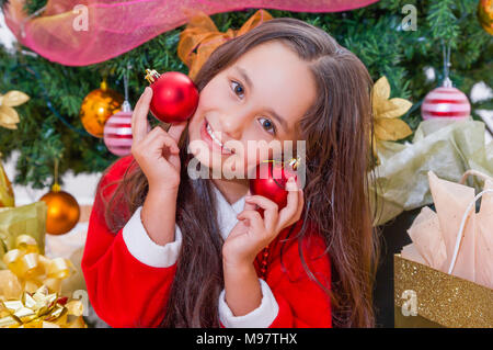 Close up of happy girl wearing a red santa costume and holding two christmas balls in her hands and pressing over her face, with a Christmas tree behind, christmas concept Stock Photo