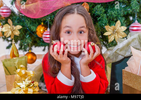 Close up of happy girl wearing a red santa costume and holding two christmas balls in her hands and pressing over her face, with a Christmas tree behind, christmas concept Stock Photo