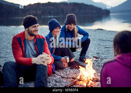 Smiling friends sitting around campfire at lakeshore Stock Photo