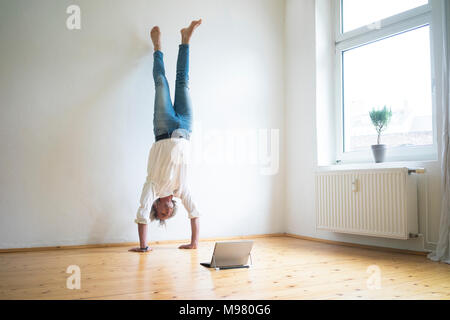 Mature man doing a handstand on floor in empty room looking at tablet Stock Photo