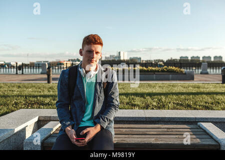 Portrait of redheaded young man sitting on bench with smartphone and headphones Stock Photo