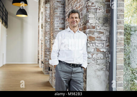 Portrait of smiling businessman leaning against brick wall in modern office Stock Photo