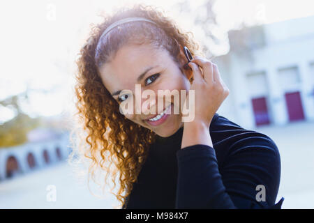 Portrait of smiling young woman outdoors putting on earphone Stock Photo