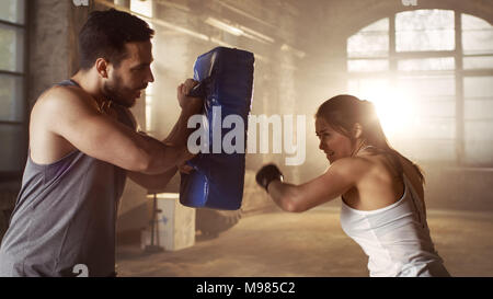 Athletic Woman Trains Her Punches on a Punching Bag that Her Partner/ Trainer Holds. She's Professional Fighter and Works out in a Hardcore Gym. Stock Photo