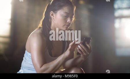 Beautiful Athletic Brunette Uses Smartphone while Resting on the Bench after Her Intensive Cross Fitness Bodybuilding Gym Training. Stock Photo