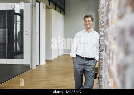 Portrait of smiling businessman leaning against brick wall in modern office Stock Photo