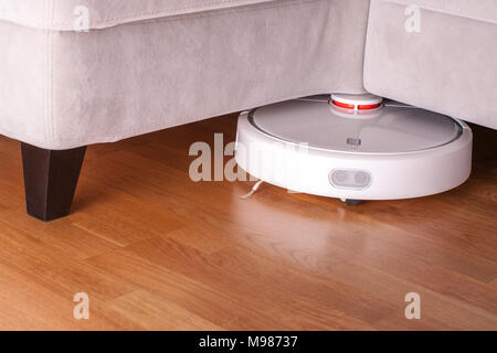 robotic vacuum cleaner runs under sofa in room on laminate floor modern smart cleaning technology housekeeping. Stock Photo
