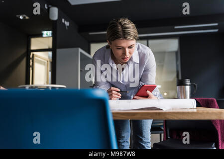 Young woman with cell phone and plan at table in office Stock Photo