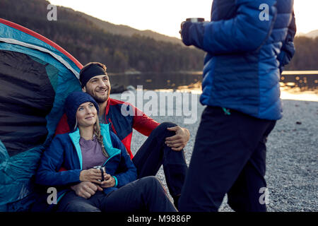 Smiling young couple with friend sitting at tent at lakeshore Stock Photo