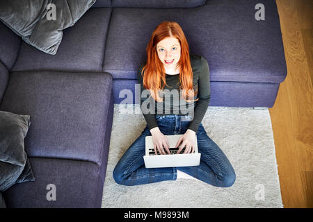 Redheaded woman sitting on floor in the living room using laptop Stock Photo