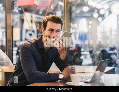 Smiling young businessman in a cafe at train station with cell phone and laptop Stock Photo
