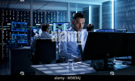 Close-up of Technical Engineer Working on His Computer With Multiple Displays in Monitoring Room. In the Background His Colleagues with Graphics Stock Photo