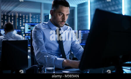 Close-up of Technical Engineer Working on His Computer With Multiple Displays in Monitoring Room. In the Background His Colleagues with Graphics Stock Photo