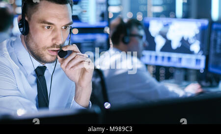 In Monitoring Room Technical Support Specialist Speaks into Headset. His Colleagues are Working in the Background. Stock Photo