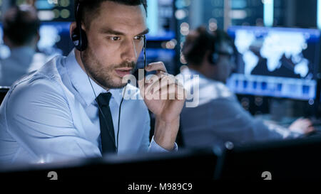 In Monitoring Room Technical Support Specialist Speaks into Headset. His Colleagues are Working in the Background. Stock Photo