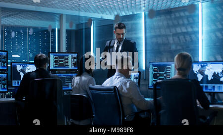 Professional IT Engineers Working in System Control Center Full of Monitors and Servers. Supervisor Holds Laptop and Holds a Briefing. Stock Photo