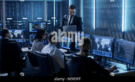 Professional IT Engineers Working in System Control Center Full of Monitors and Servers. Supervisor Holds Laptop and Holds a Briefing. Stock Photo